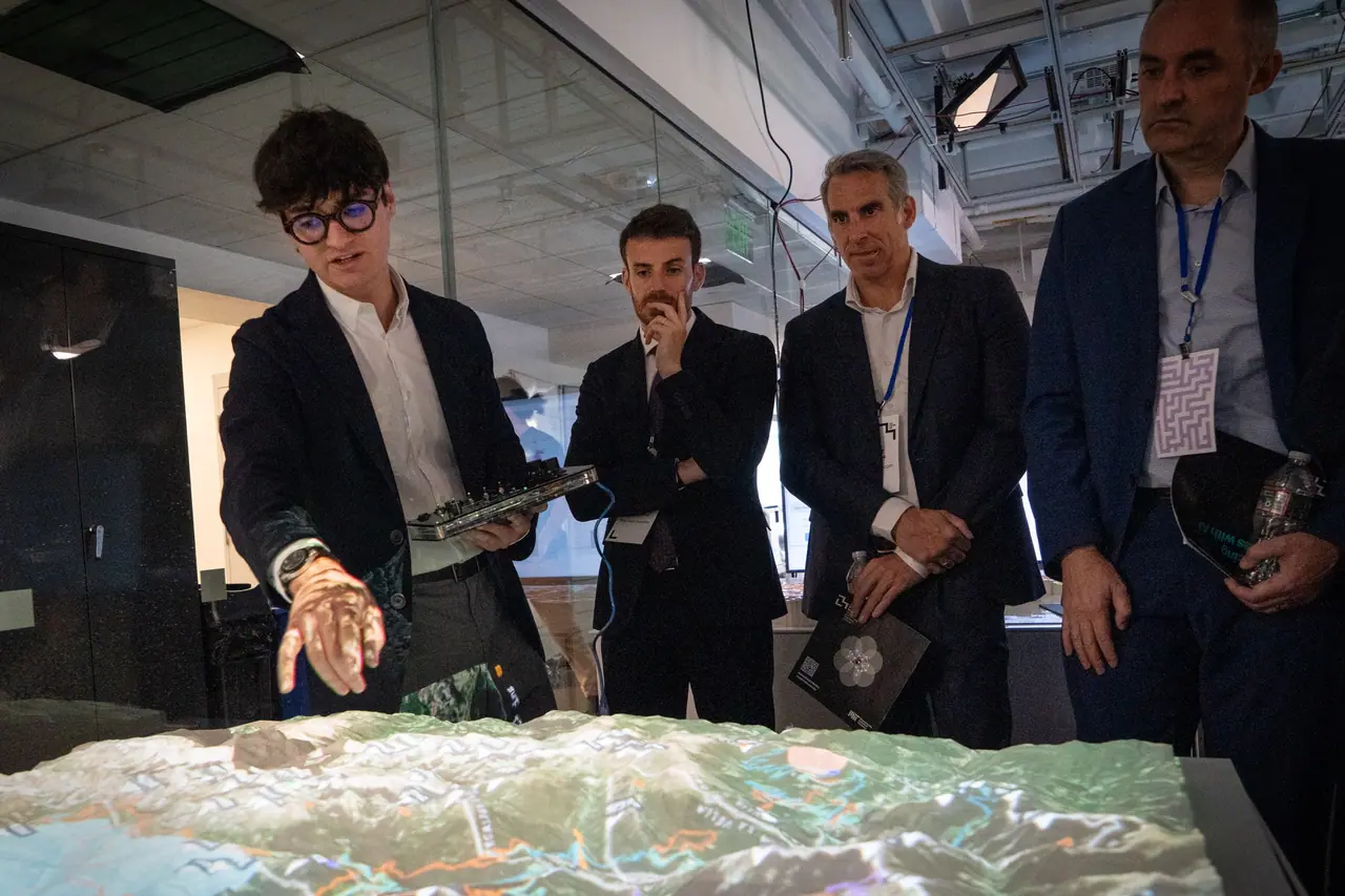Marcel, in a dark blazer and round black glasses, holds the hand-built control panel in one hand and reaches toward the projection-lit relief model of Andorra while three members of the Andorran delegation look on.