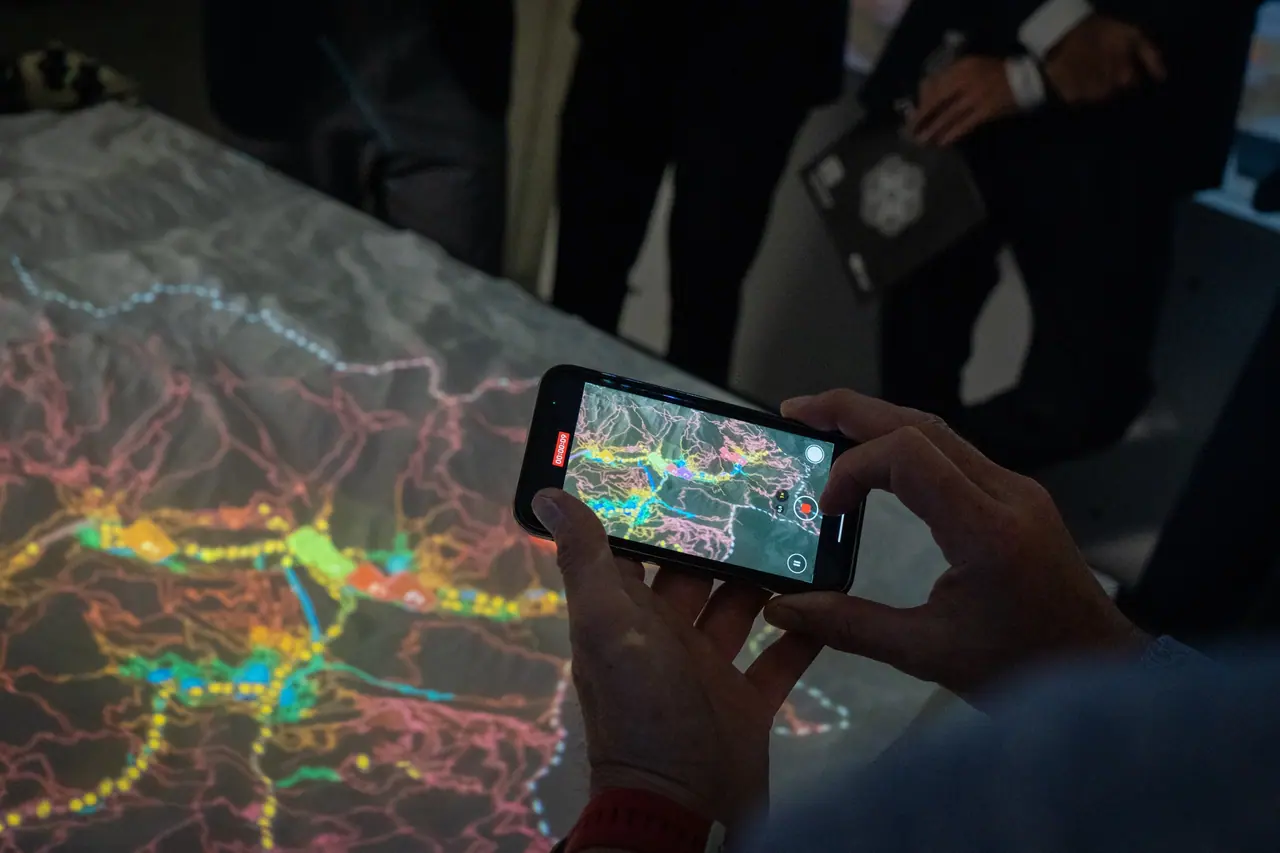 A visitor holds up a phone to film the projection-lit terrain model; the road network glows pink, orange and yellow on the table beside the phone screen.
