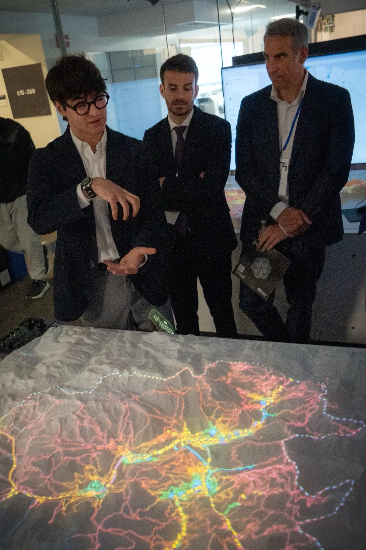 Marcel talks with his hands beside the terrain model while two men in dark suits listen; on the table the road and trail network of Andorra glows in orange, yellow and green.