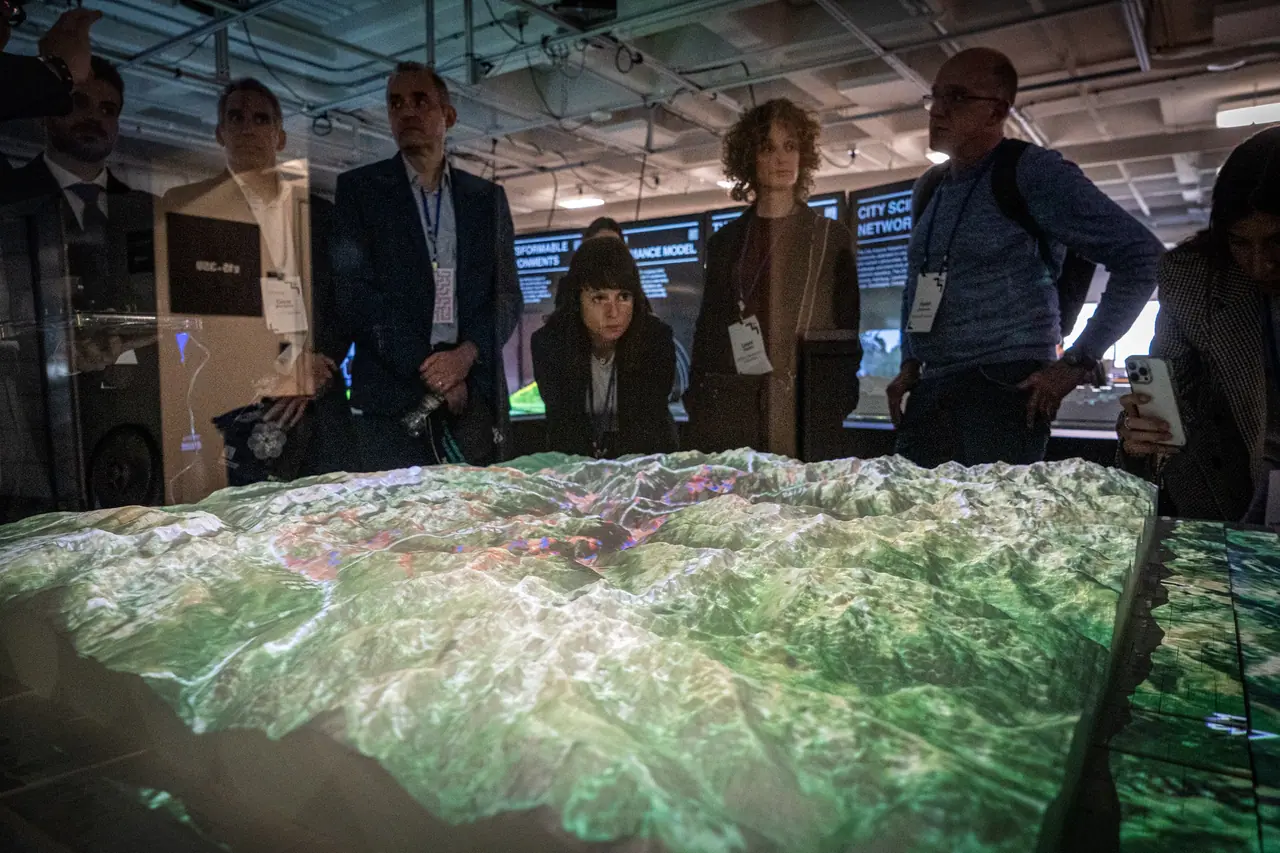 Visitors wearing lanyards stand behind the relief model of Andorra, its mountains lit green by the projector; a woman leans in to look closer.