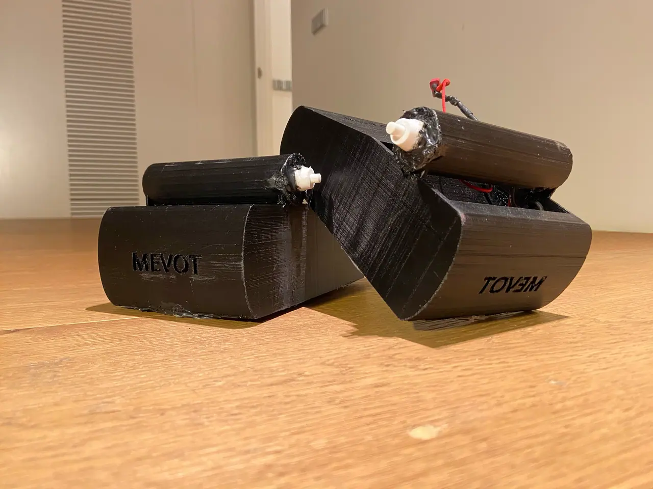 Two black 3D-printed cases lettered MEVOT on a wooden floor, each carrying a short cylinder with a white button; one case is tilted open at an angle with a red wire showing.