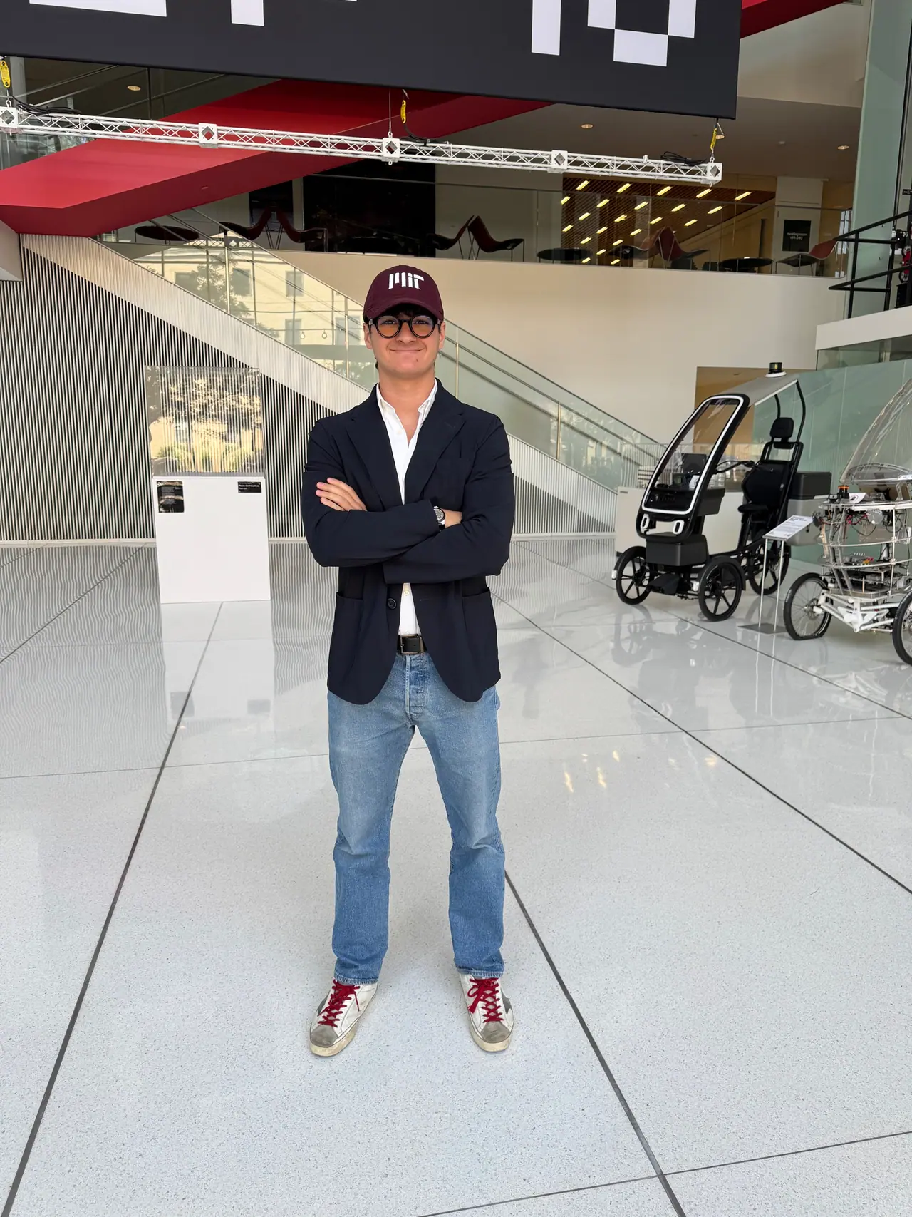 Marcel in round glasses and an MIT cap, arms crossed, standing on a polished floor in a bright atrium with a glass balcony behind him and a small electric city vehicle at the right edge.