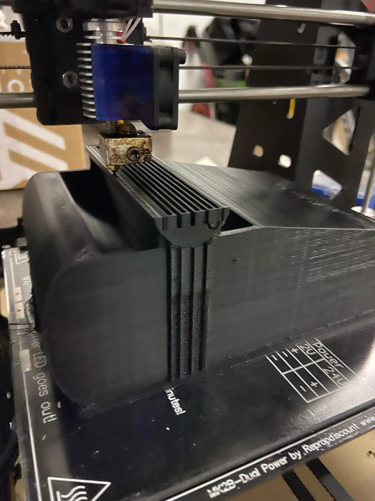 Close view of a desktop 3D printer's hot end above a large black printed part, a rounded box with a ribbed edge, on a heated bed printed with the words MK2B Dual Power.