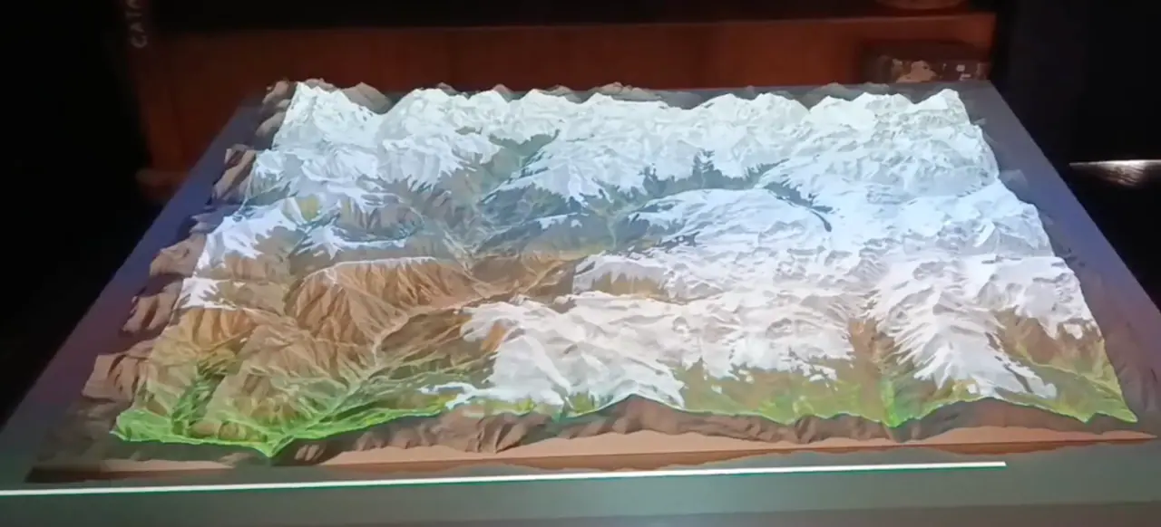 A physical relief model of Andorra's mountains lit by a projector in a dark room: white peaks, brown slopes and green valley floors, seen at an angle from one corner of the table.